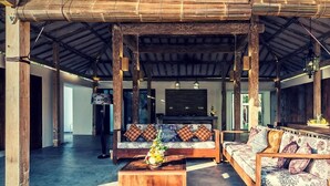 A spacious open-plan living area with a high ceiling and exposed wooden beams. There are two sofas with patterned cushions and a wooden coffee table with a vase of flowers.