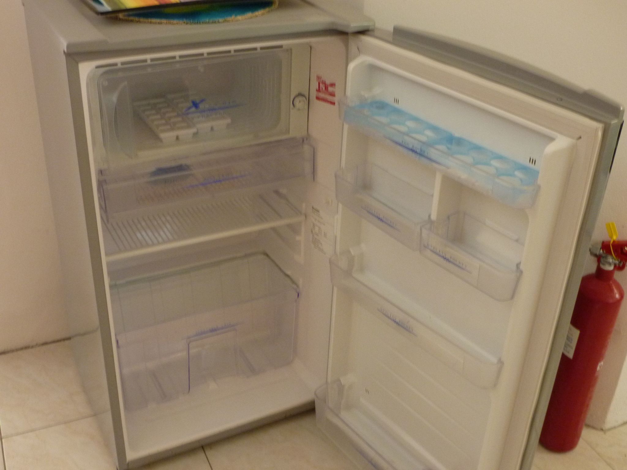 Et åbent køleskab med flere hylder og rum, en brandslukker ophængt på væggen, og en blå bakke ovenpå.