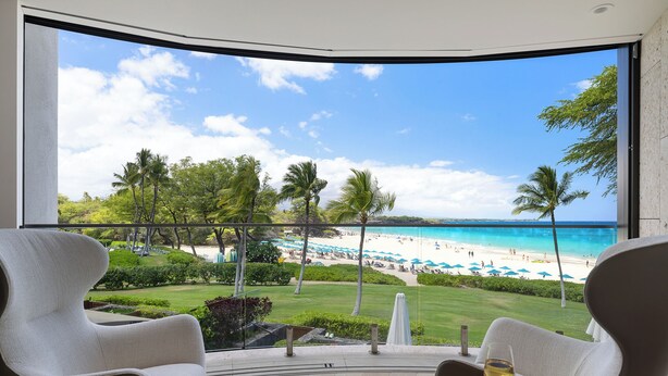 A balcony with two chairs and a glass table with a wine glass, overlooking a beach with palm trees and a clear blue sky.