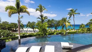 A poolside area with lounge chairs, a clear view of the ocean, and palm trees.