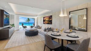 A modern living room with a large window overlooking the ocean, a dining area with a round table, and a kitchen area with a large mirror.