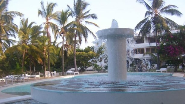 Una piscina con una fuente central rodeada de sillas de descanso y palmeras.