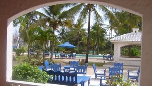 Un área de estar al aire libre con sillas y mesas azules, rodeada de palmeras y una piscina.