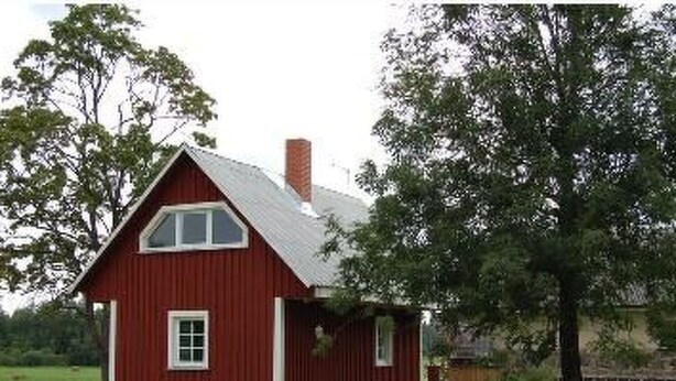 Ein rotes Holzhaus mit einem Satteldach und Schornstein, umgeben von einer grünen Wiese und Bäumen.