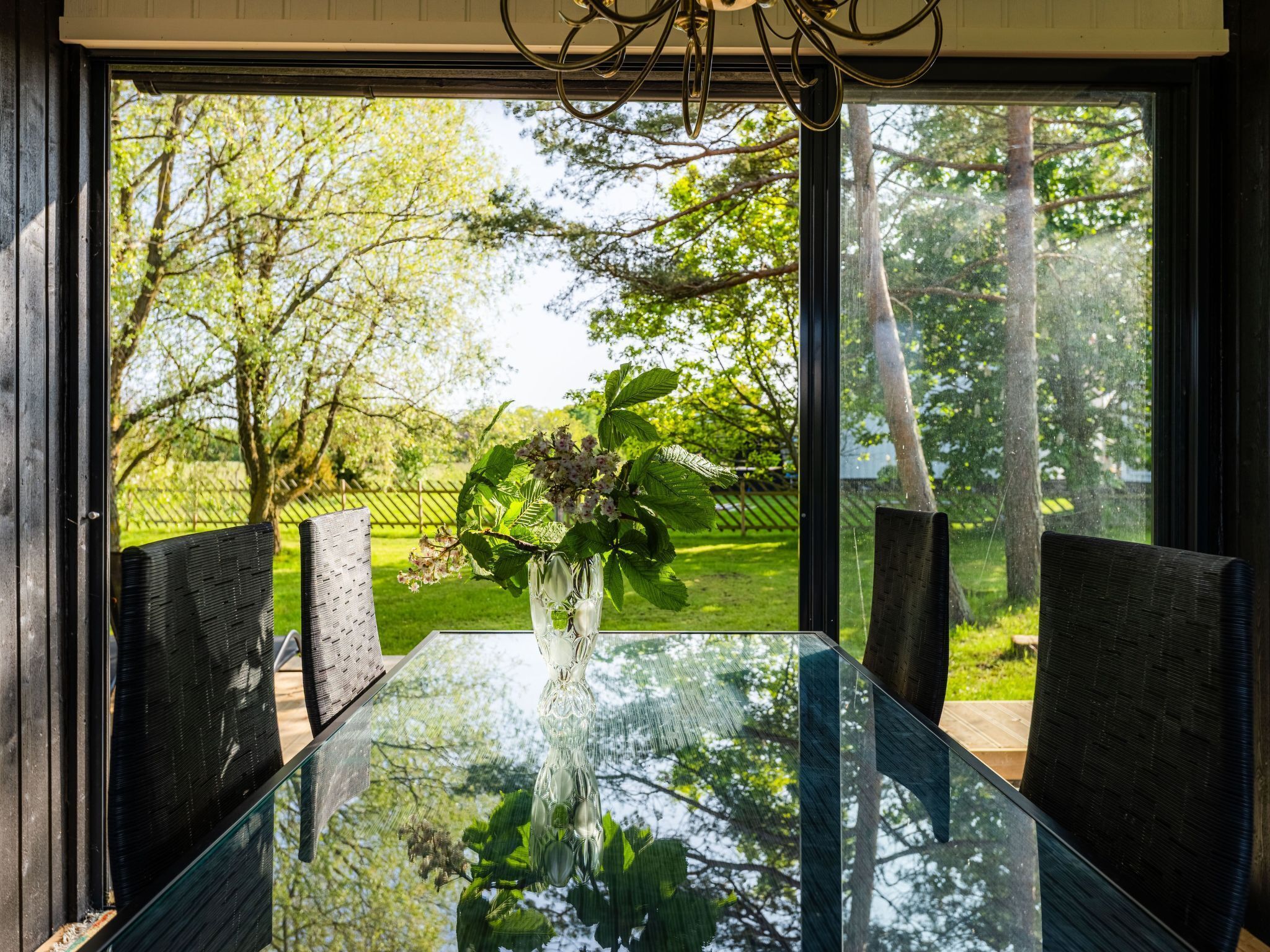 Una mesa de comedor con cubierta de vidrio, rodeada de sillas, frente a un ventanal con vista a un jardín y árboles.