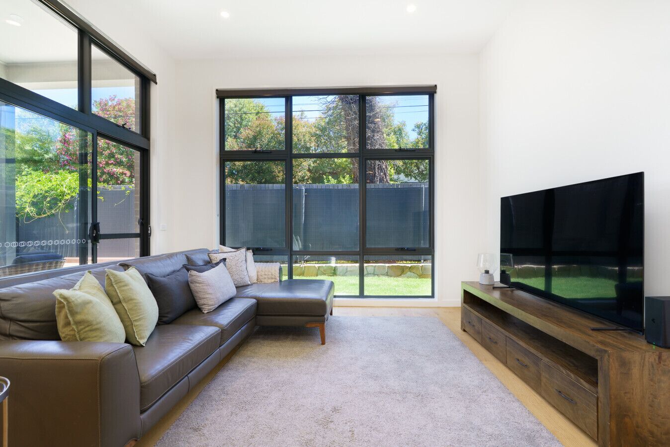 A modern living room with a sectional sofa, a flat-screen TV, and large windows offering a view of a backyard.