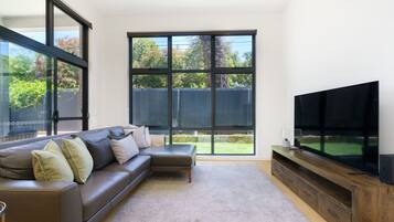A modern living room with a sectional sofa, a flat-screen TV, and large windows offering a view of a backyard.