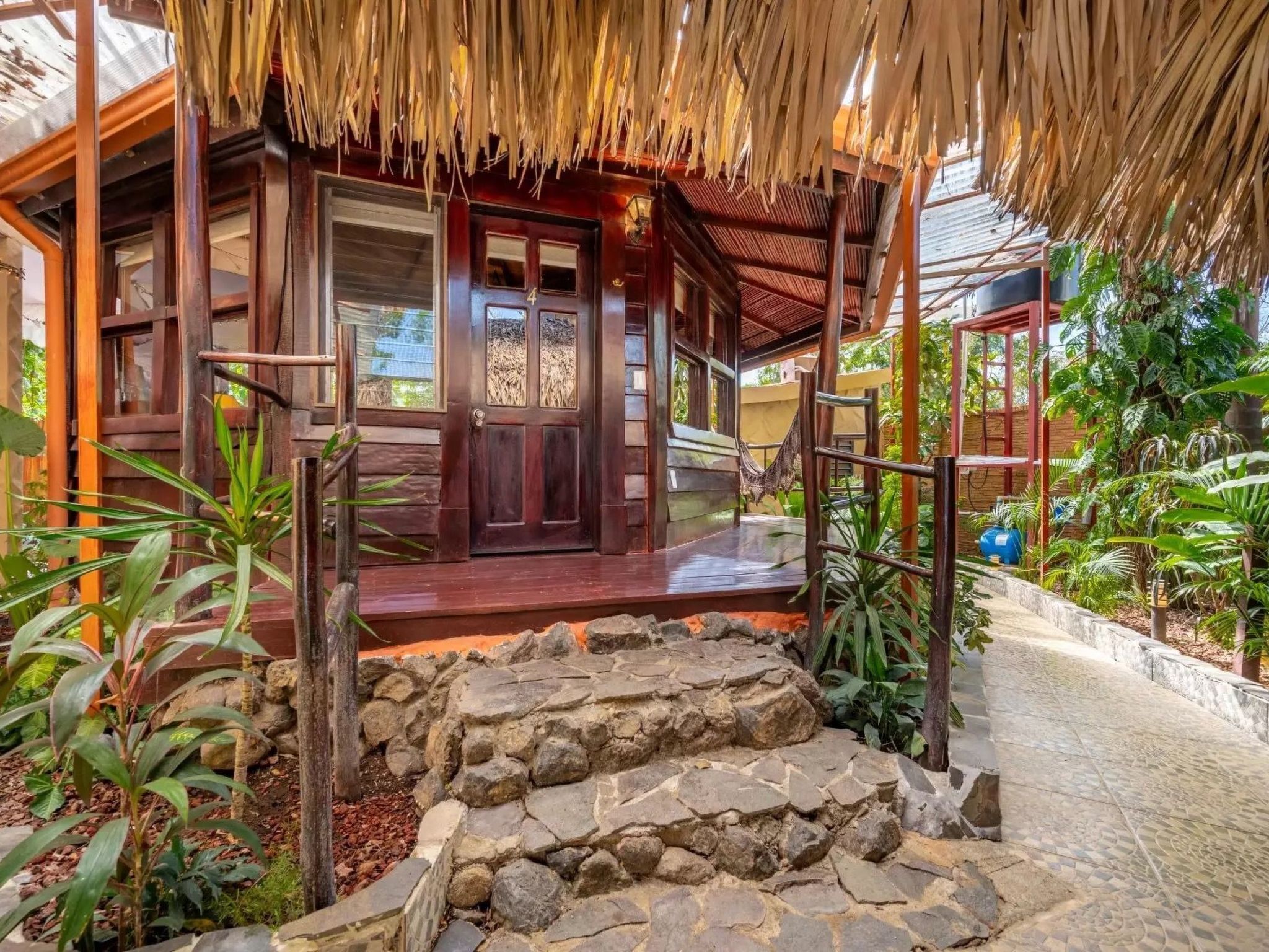 Una casa de madera con techo de paja, una escalera de piedra y un jardín con plantas tropicales.