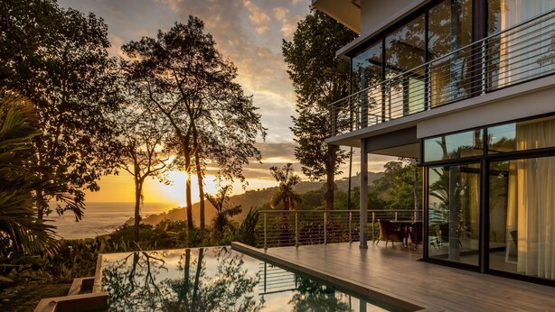 Ein moderner Poolbereich im Freien mit einer Holzterrasse, umgeben von üppiger Vegetation und mit Blick auf eine malerische Meereslandschaft bei Sonnenuntergang.