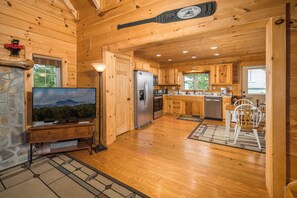 Interior de una cabaña con chimenea de piedra, televisor, cocina con armarios de madera y comedor.