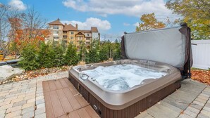 Un bain à remous dans un jardin, avec une terrasse en bois, entouré d’arbres et avec un bâtiment visible au loin.