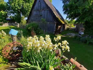 Um estábulo rústico com telhado de colmo, rodeado por vegetação exuberante e diversas flores.