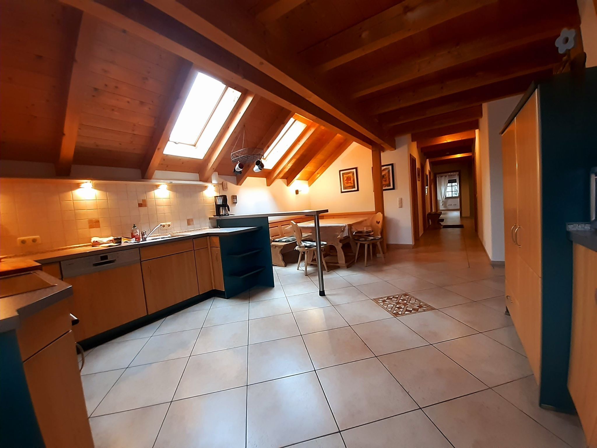 Una cocina con gabinetes de madera, un comedor con mesa y sillas, y un techo inclinado con tragaluces.