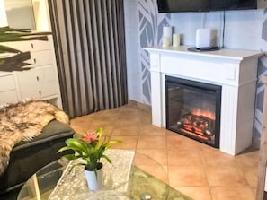 En moderne stue med pejs, et glasbord med en plante, et fuskøde og et tv ophængt på væggen.