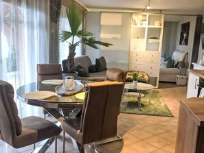 Ein modernes Wohnzimmer mit einem Glastisch, bequemen Sitzgelegenheiten, einer großen Kübelpflanze und einer Küchenzeile im Hintergrund.