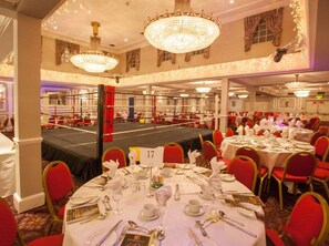 Ein Festsaal mit runden Esstischen, einer Boxarena in der Mitte und Kronleuchtern an der Decke.