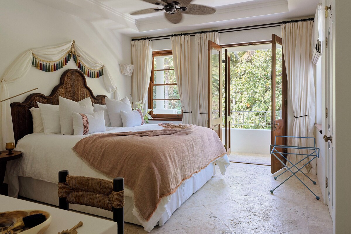 A bedroom with a large bed, a ceiling fan, a window with curtains, and a view of greenery outside.