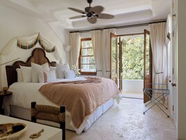 A bedroom with a large bed, a ceiling fan, a window with curtains, and a view of greenery outside.