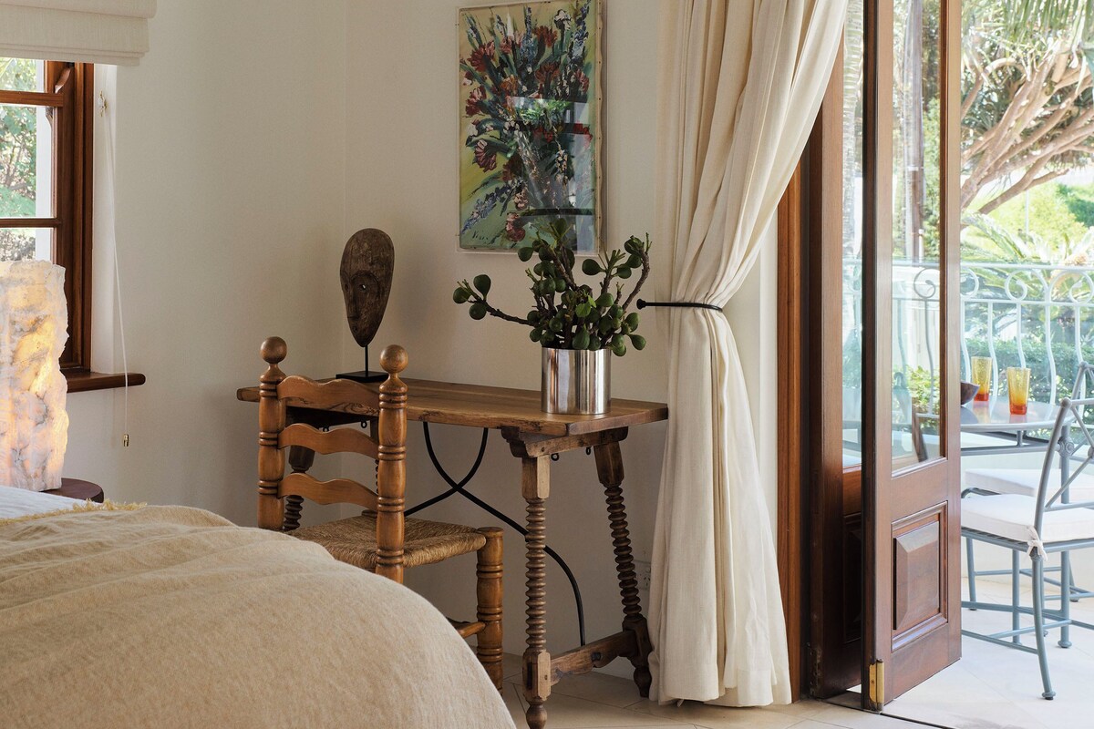 A bedroom with a bed, a wooden chair, a small table with a vase of flowers, a painting on the wall, and a view of the outdoors through a door.