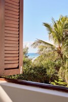 A window with wooden shutters, a view of a beach, and palm trees.