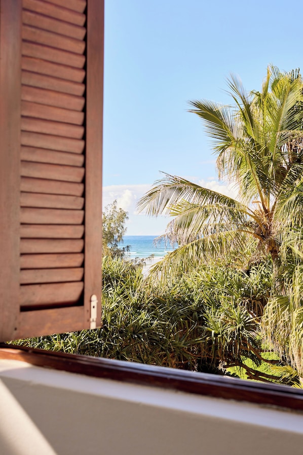A window with wooden shutters, a view of a beach, and palm trees. - Raes on Wategos (Byron Bay)