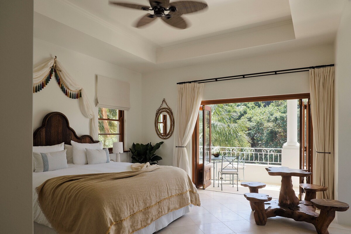 A bedroom with a large bed, a ceiling fan, a balcony with a table and chairs, and a view of greenery outside.