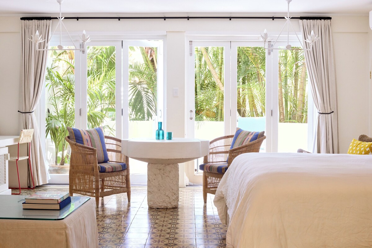 A hotel room with a bed, two wicker chairs, a small table, and a view of palm trees through large windows.