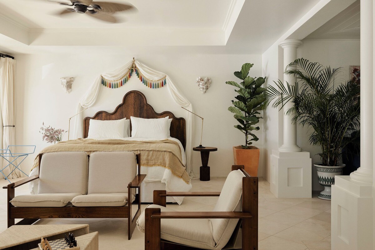 A bedroom with a large bed, two armchairs, a side table, and a ceiling fan.