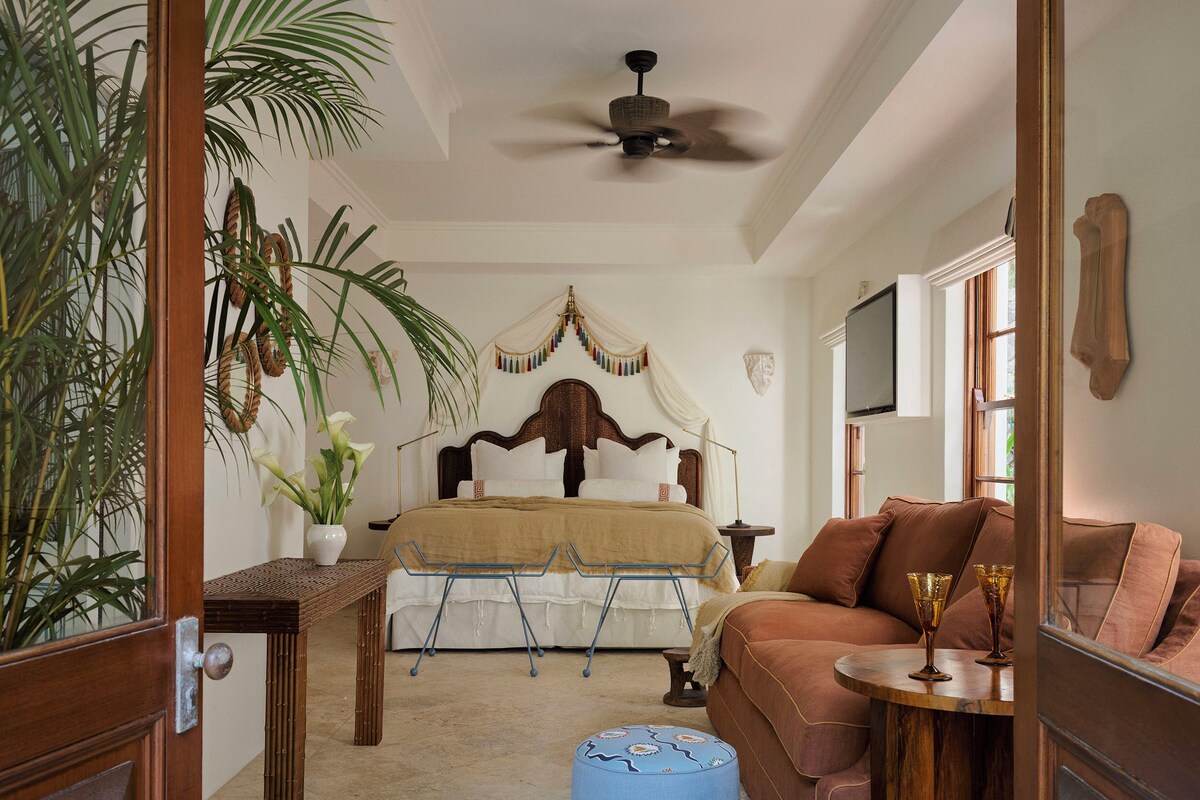 A bedroom with a bed, a wooden headboard, a brown sofa, two ottomans, a ceiling fan, and a window.