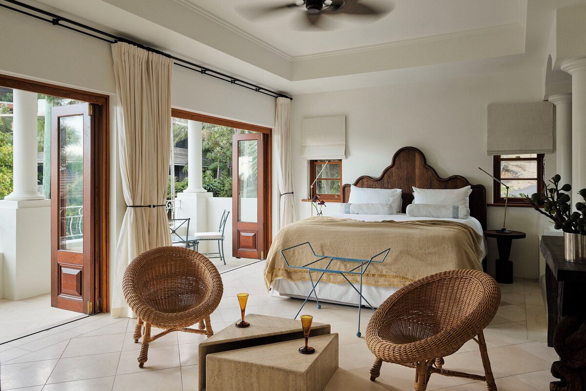 A bedroom with a bed, two wicker chairs, a small table, and a ceiling fan.
