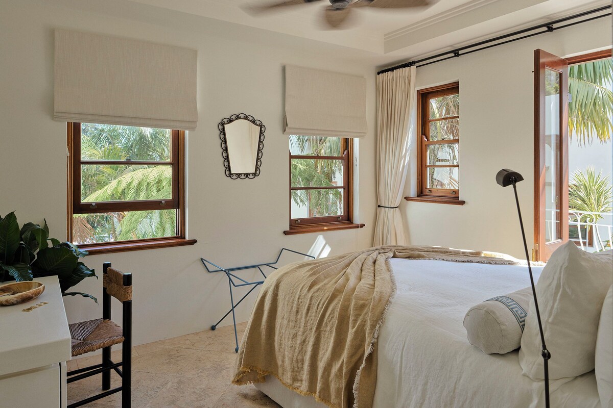 A bedroom with a bed, a ceiling fan, two windows with curtains, a chair, and a desk.