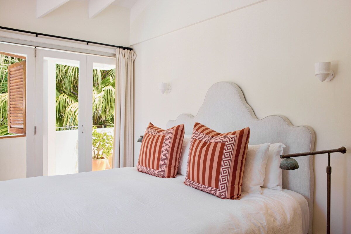 A hotel room with a bed, two pillows, a nightstand, a lamp, and a view of the outdoors.
