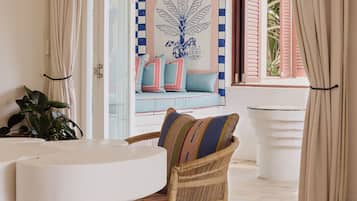 Quarto com cadeira de vime, sofá branco com almofadas estampadas, mesa branca e vista para o exterior através de uma porta aberta.