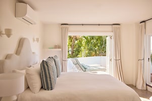 A bedroom with a bed, pillows, a nightstand, a lamp, and a view of a balcony with outdoor furniture.