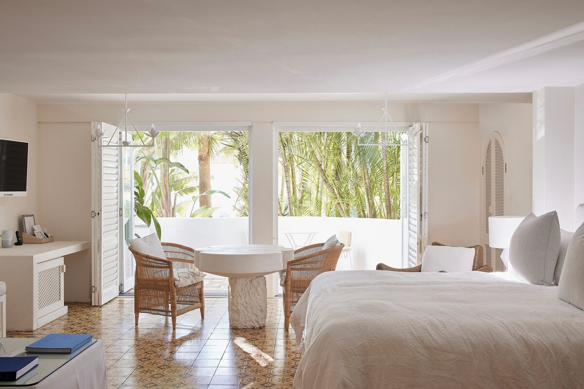 A hotel room with a bed, a desk, a chair, a table, and a large window with a view of palm trees.