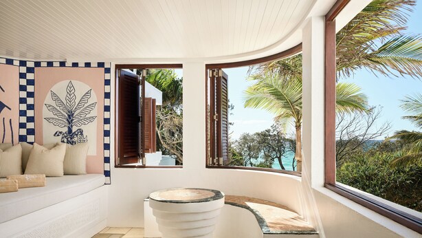 A modern bathroom with a toilet, a bench, and a window offering a view of palm trees and the ocean. - Raes on Wategos  (Byron Bay)