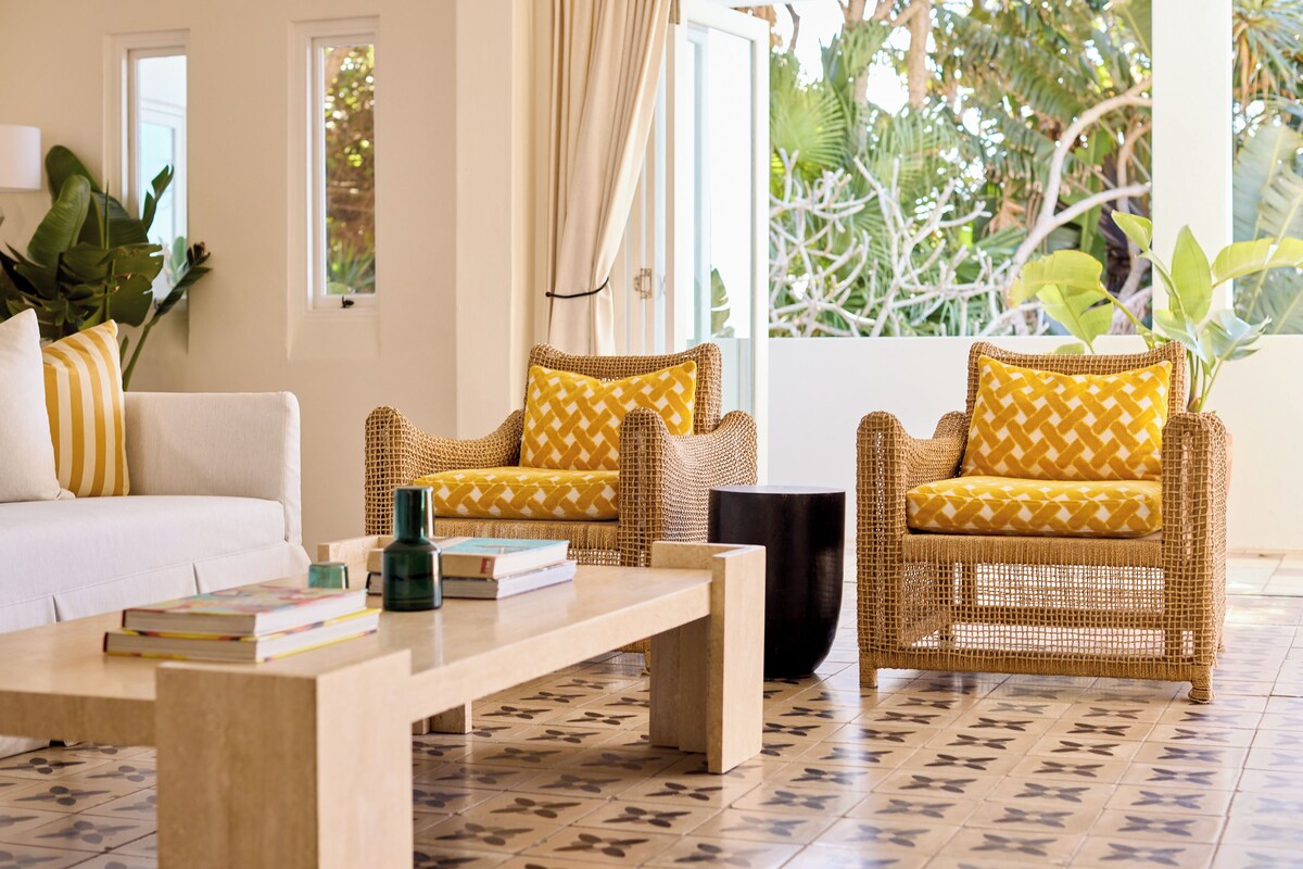 A room with a sofa, two wicker chairs with yellow cushions, a wooden coffee table, and a view of greenery outside.