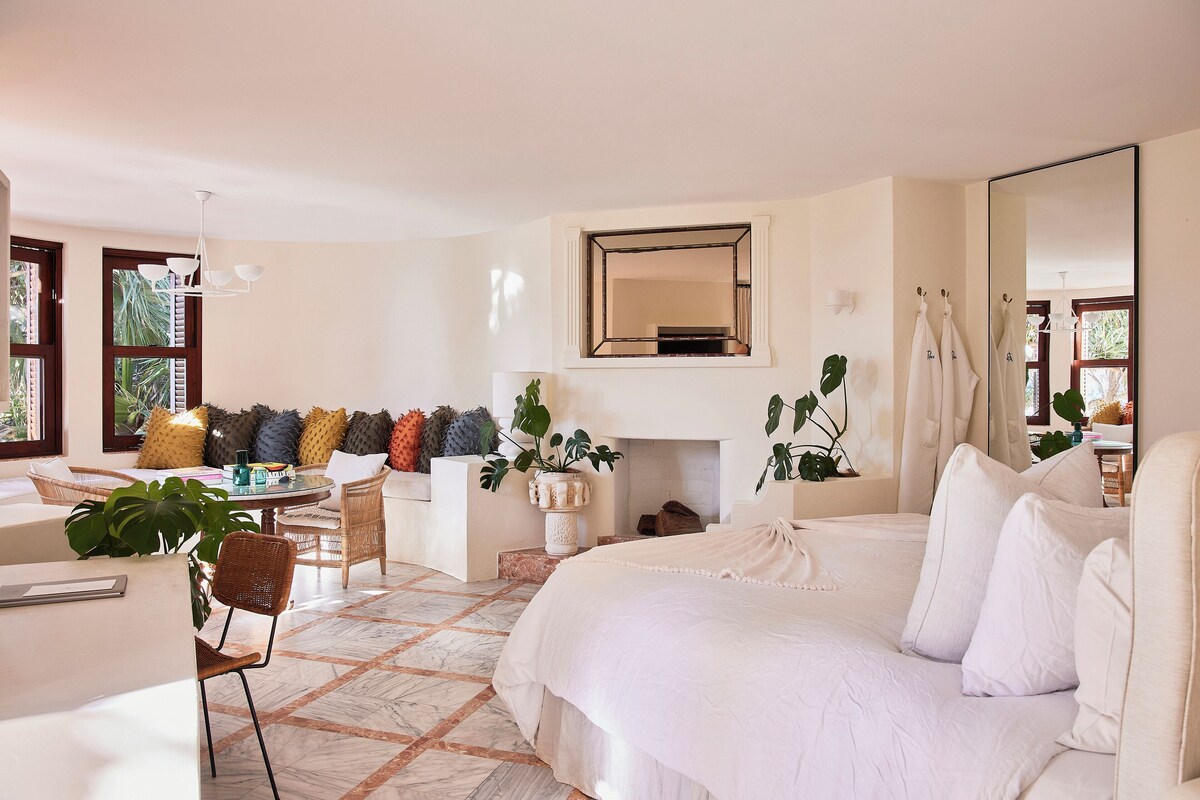 A bedroom with a large bed, a sitting area with a table and chairs, a fireplace, and a large mirror.