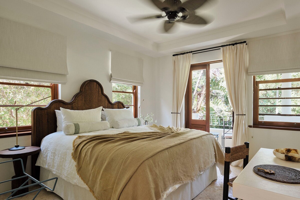 A bedroom with a bed, a ceiling fan, a window with curtains, and a table with a bowl and a mat.