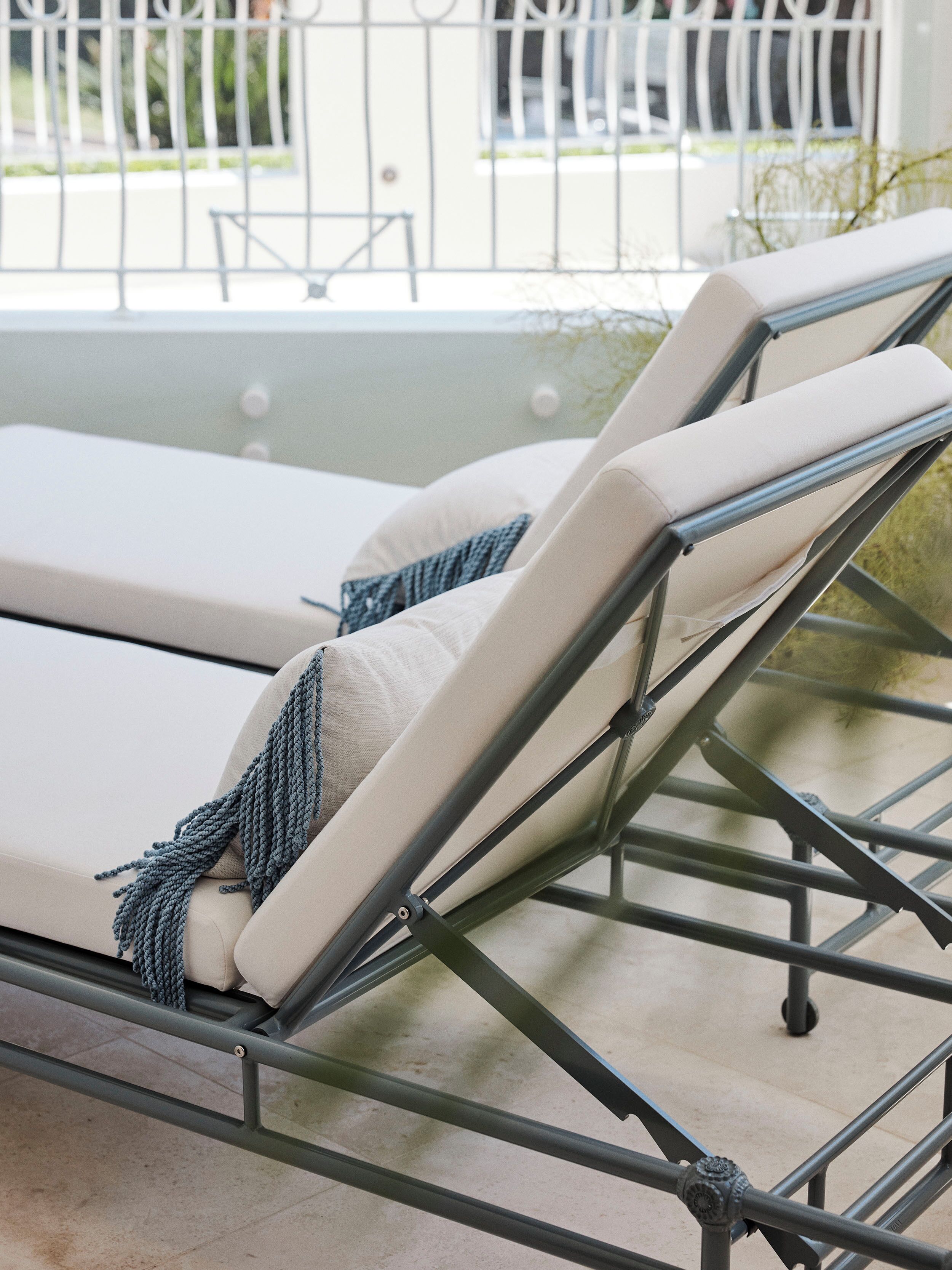 A sun lounger with a metal frame and beige cushions, positioned on a balcony with a view of a white fence and greenery.