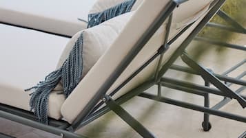 A sun lounger with a metal frame and beige cushions, positioned on a balcony with a view of a white fence and greenery.
