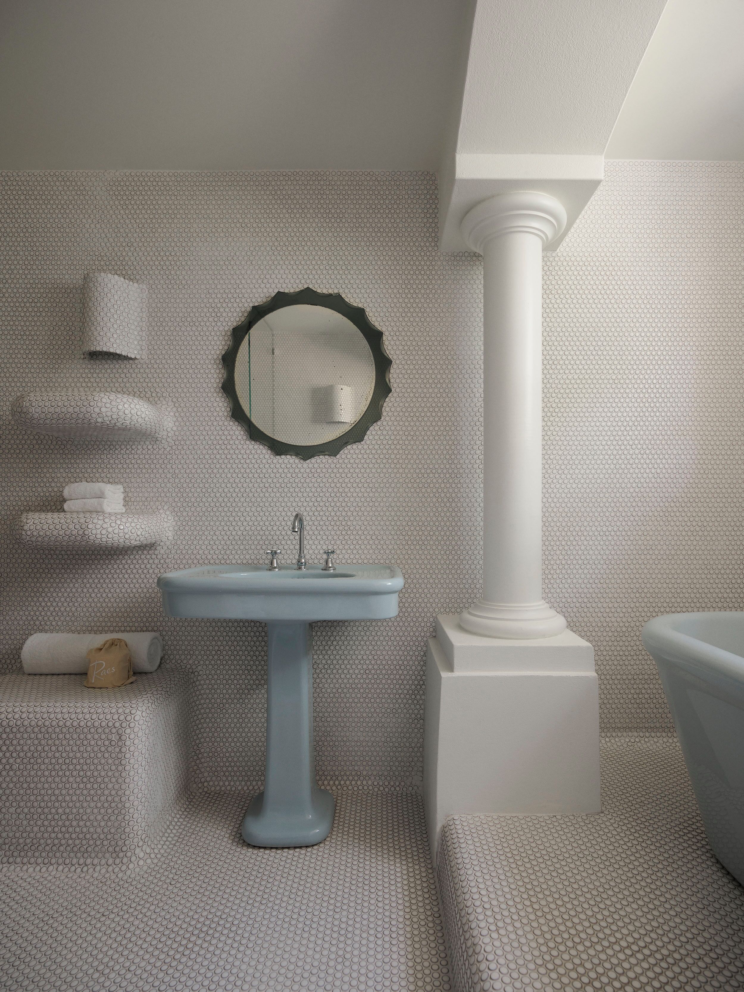 A bathroom with a pedestal sink, a round mirror, a white bathtub, and a towel rack with towels.