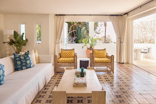 A room with a white sofa, two wicker chairs with yellow cushions, a coffee table with books, and a view of tropical plants through large windows. - Raes on Wategos (Byron Bay)