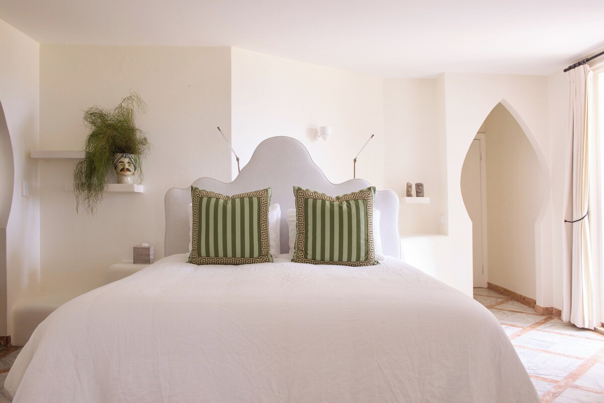 A bedroom with a large bed, two pillows with a geometric pattern, a wall-mounted plant, and a door leading to another room.