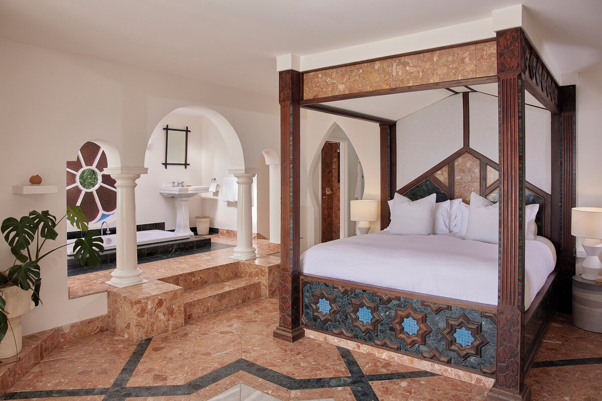 A four-poster bed with a geometric-patterned footboard, a marble-tiled floor, and a white sink with a mirror above it.