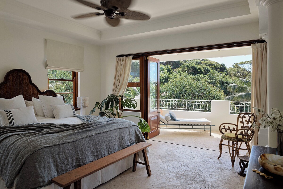 A bedroom with a large bed, a ceiling fan, a balcony with a view of greenery, and a chair.