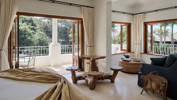 A spacious room with a large bed, a wooden table, and a blue sofa. There are large windows with curtains, a balcony with a railing, and a view of trees and a building. - Raes on Wategos  (Byron Bay)