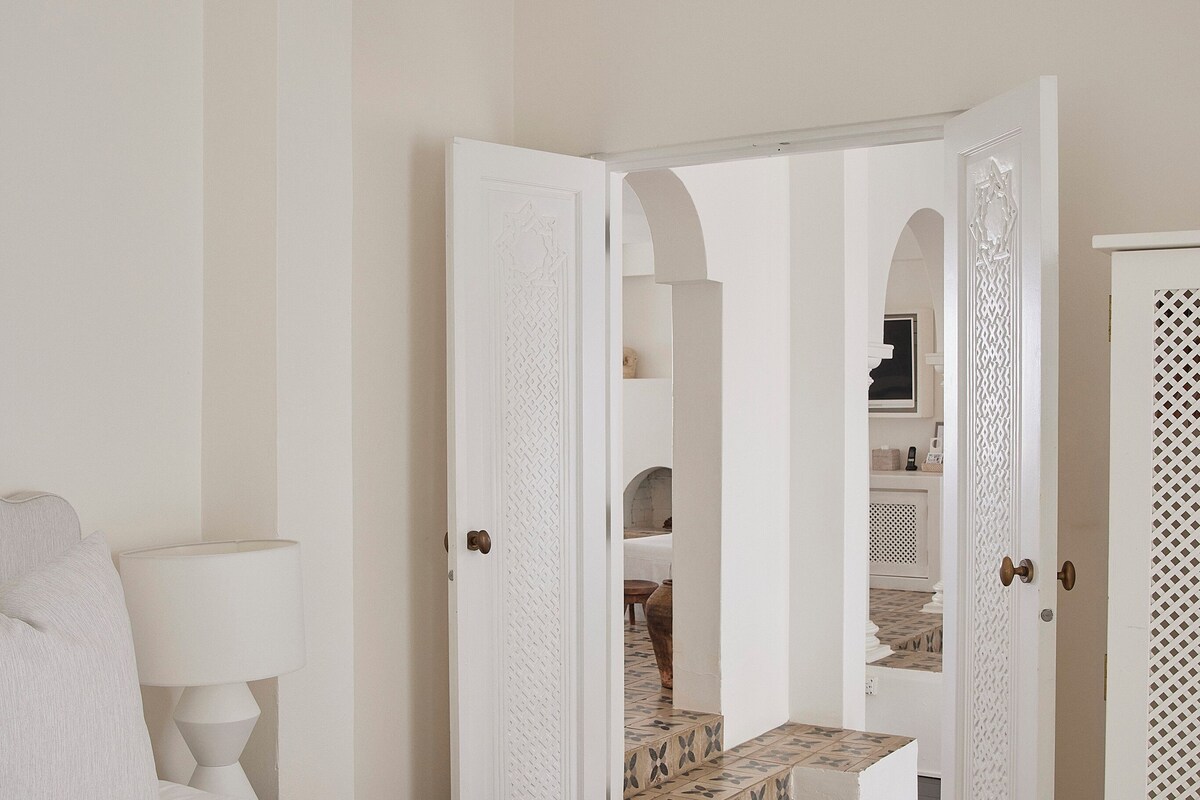 A bedroom with a large bed, a nightstand, and a lamp. There are two doors leading to other rooms.