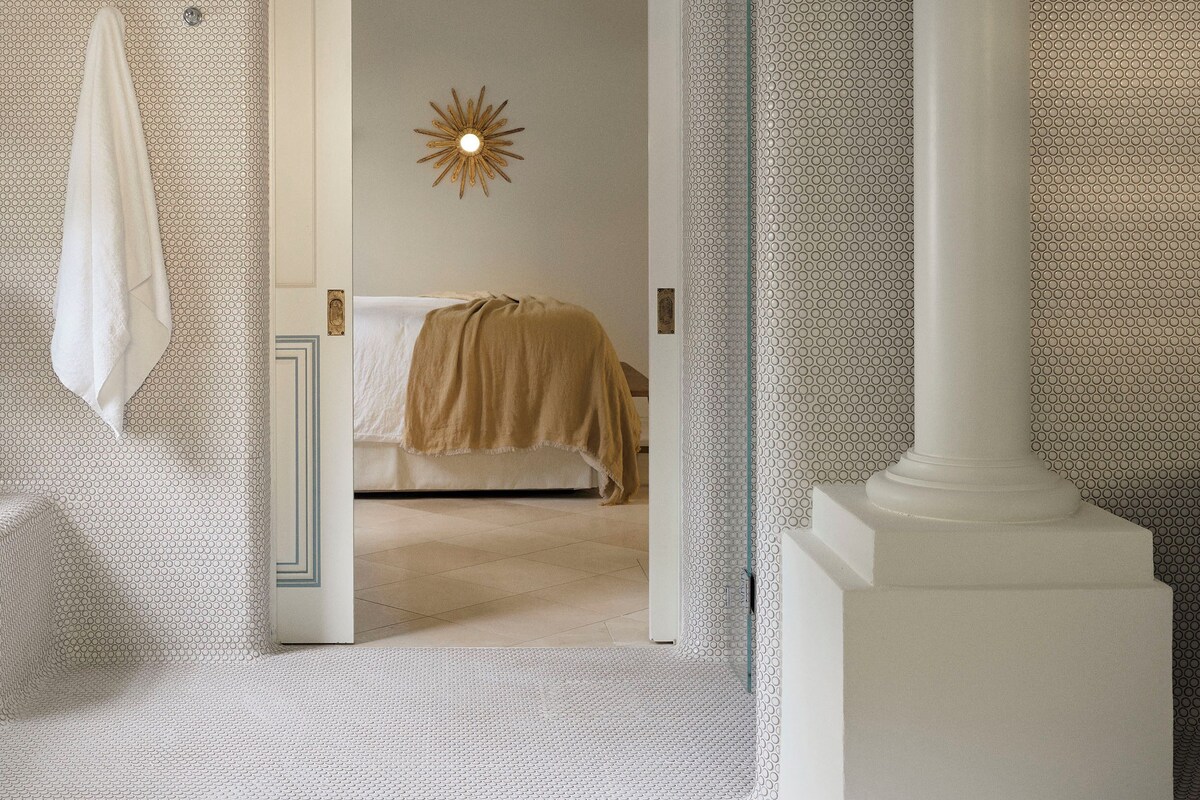 A bedroom with a patterned wallpaper, a bed with a beige blanket, a ceiling fan, and a sun-shaped light fixture.