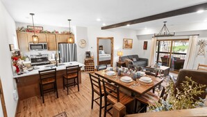 A modern kitchen with wooden cabinets, a microwave, and a refrigerator. There is a dining area with a table set for four, surrounded by wooden chairs. A sofa and a lamp are visible in the background. - Tall Timbers Cabin, Private Hot Tub, Outdoor Pool, Free Daily Tickets & Near Thunder Ridge (Hollister)